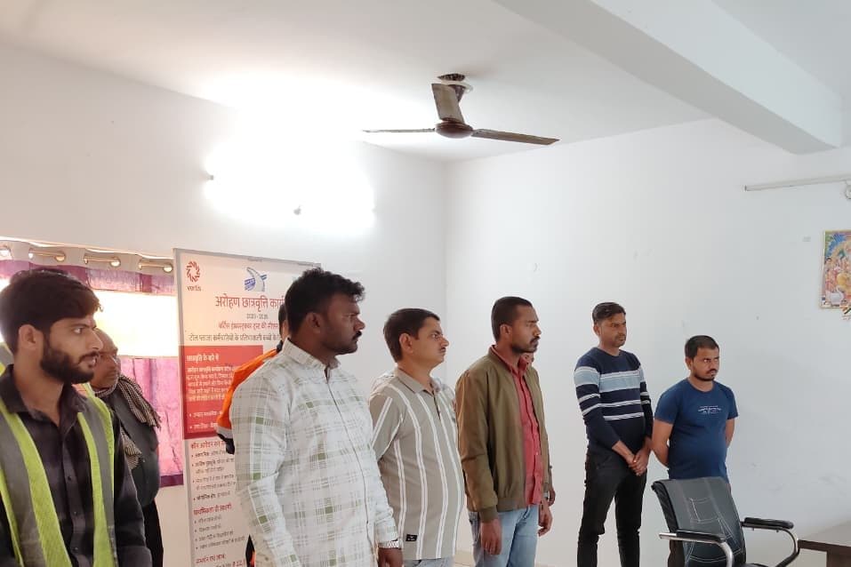 A group of men standing and listening to a presentation, beside a printed Aarohan scholarship programme banner in a room with ceiling fans